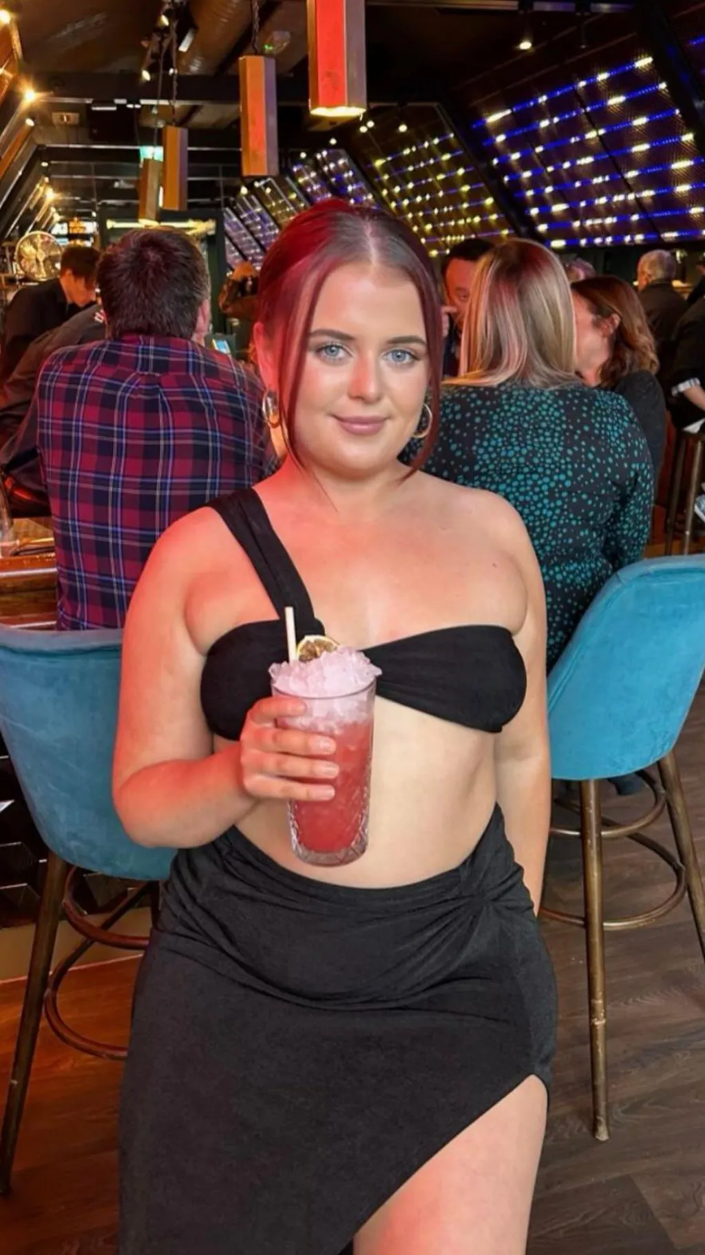 Katie wearing a black dress while she holds a cocktail in her hand. The drink is pink and has a straw. Behind her are other people in the bar sitting on chairs.