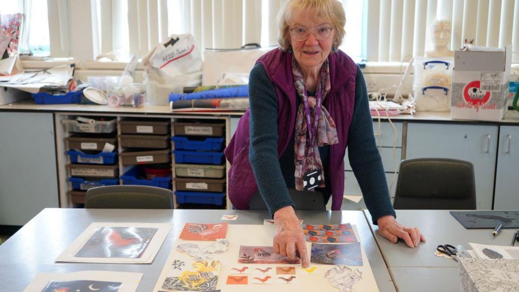 Heather Monaghan is pictured in a blue top and fuchsia gilet in a classroom pointing towards her art work which depicts cockerels  