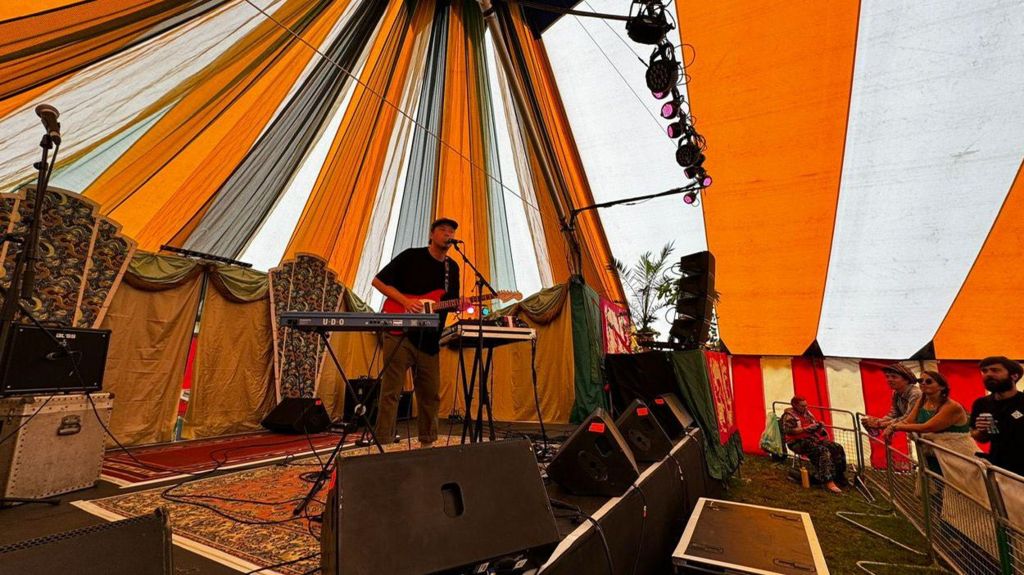Max Rad on stage playing guitar and singing into a microphone in a striped tent