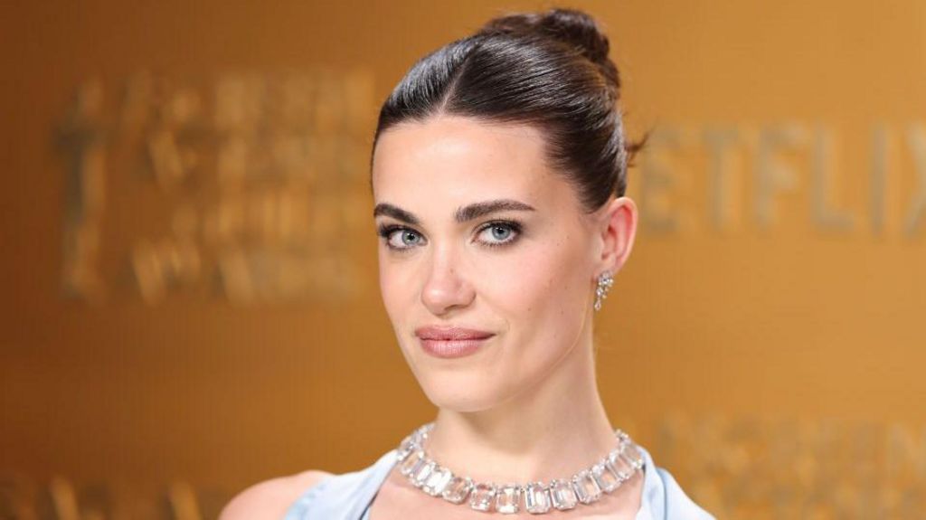 Genevieve Chenneour is seen with dark hair styled in a sleek bun and is photographed in front of a gold backdrop with Netflix branding. She is wearing a light blue satin gown, a diamond necklace and matching earrings, and is looking directly at the camera with a slight smile.