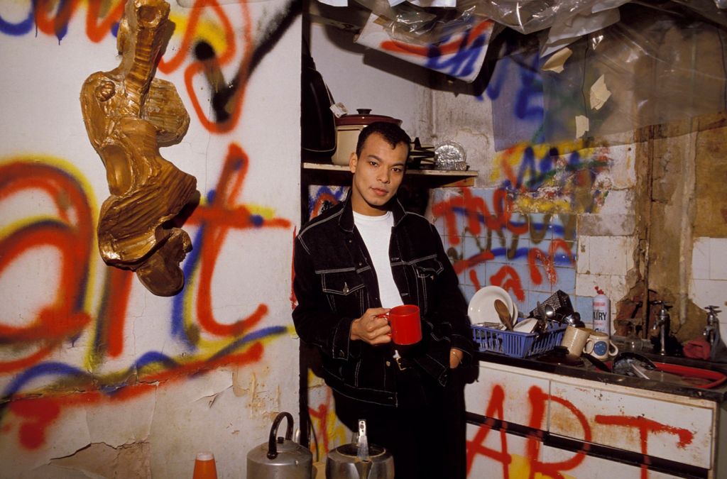 Roland Gift poses for a photograph while leaning against the wall of a dirty, delapidated kitchen with graffiti all over it and holds a mug while looking at the camera wearing a denim top, taken in 1980.