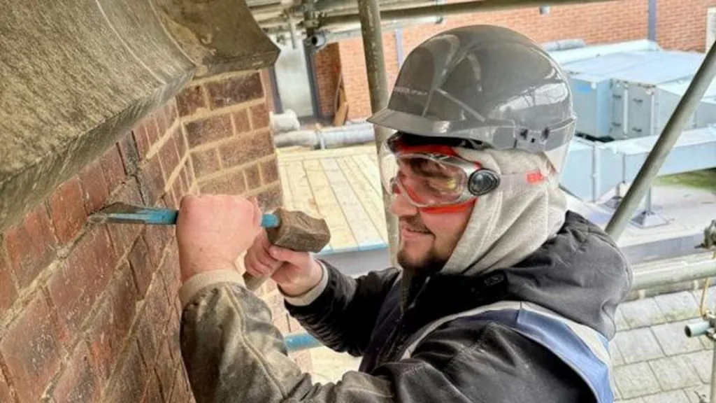 A man in a black dusty jacket with a grey helmet and eye goggles chiselling at a brick wall. 