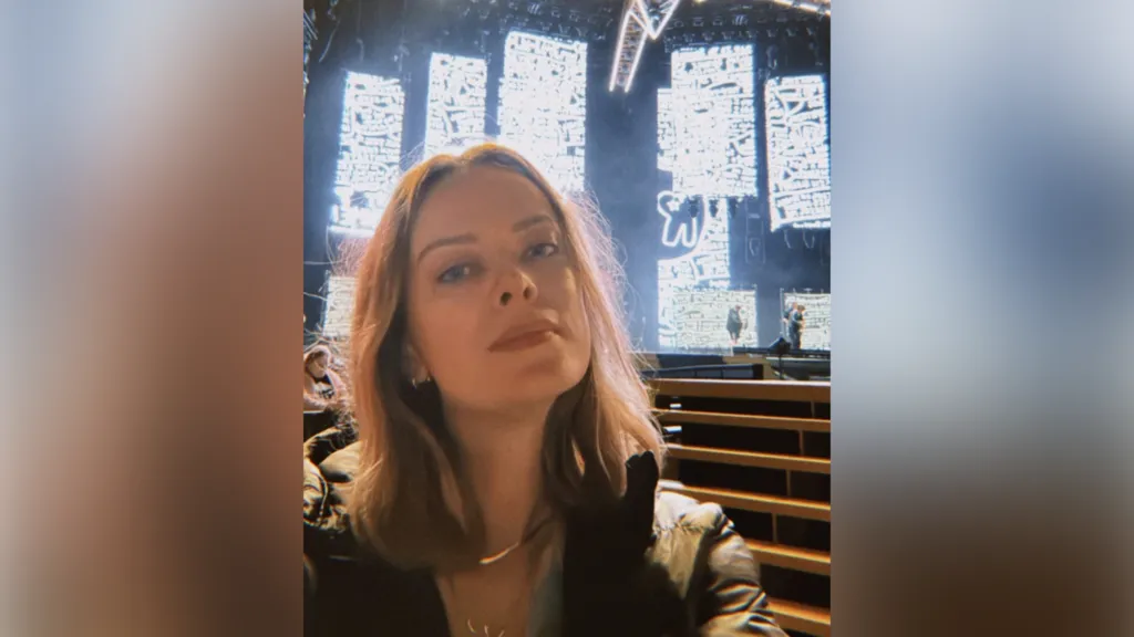 A woman with dark shoulder-length hair takes a selfie in front of a stage which has black and white projections on it.