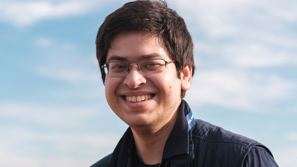 A young man in glasses smiles widely against a cloudy blue sky. The sunlight hits the side of his face, suggesting a late autumn afternoon. 