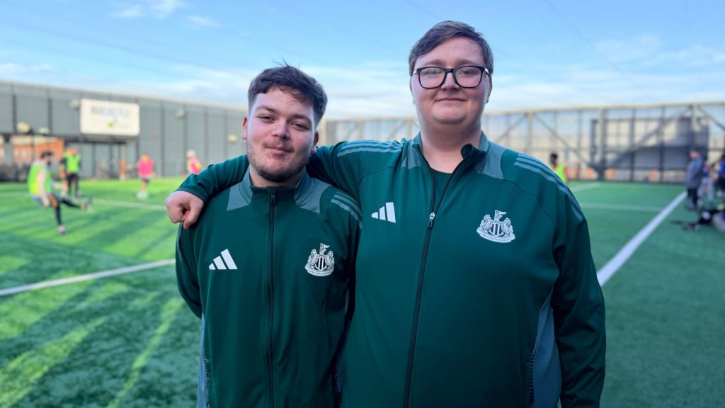 Two young men wearing Newcastle United Football Club-branded tracksuit tops. They are standing in front of a football pitch where a game is under way.