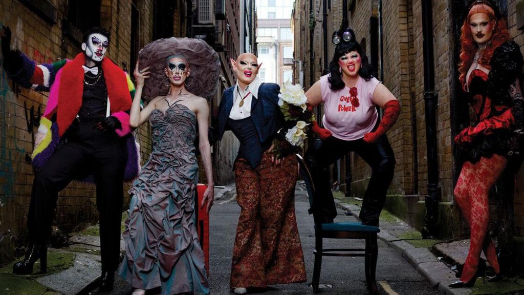 Five drag performers pose in an alley. The first, dressed as a man wears a white face and multi-coloured coat. The second has a grey dress with a large round hate. The third wears a blazer and wide trouers, the third stands on a chair in a t-shirt and red elbow-length gloves. The last rests with her back on the wall wearing red patterned tights and a corset.