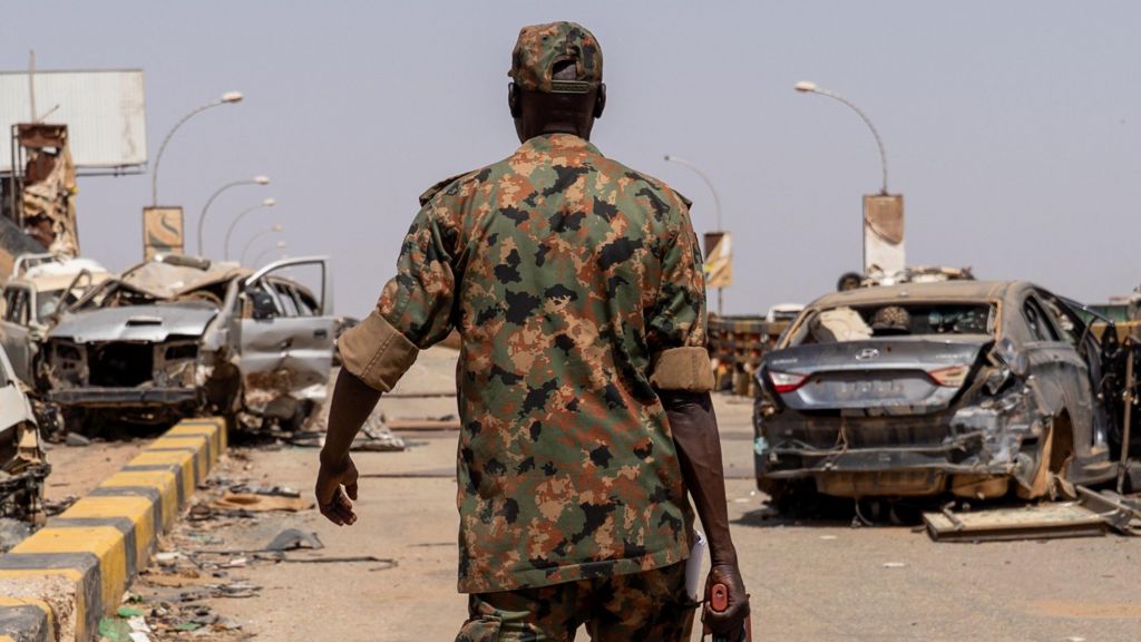 A soldier in military uniform and holding a gun, walks on a street, with several damaged cars in front of him