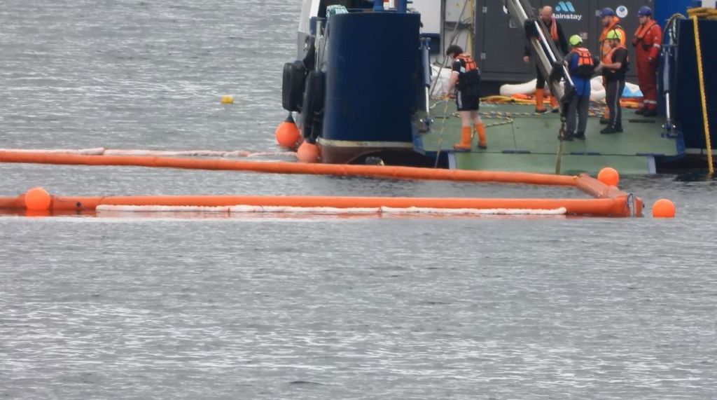 Red floating bollards with a boat moored alongside with workers aboard observing and working around the spill site