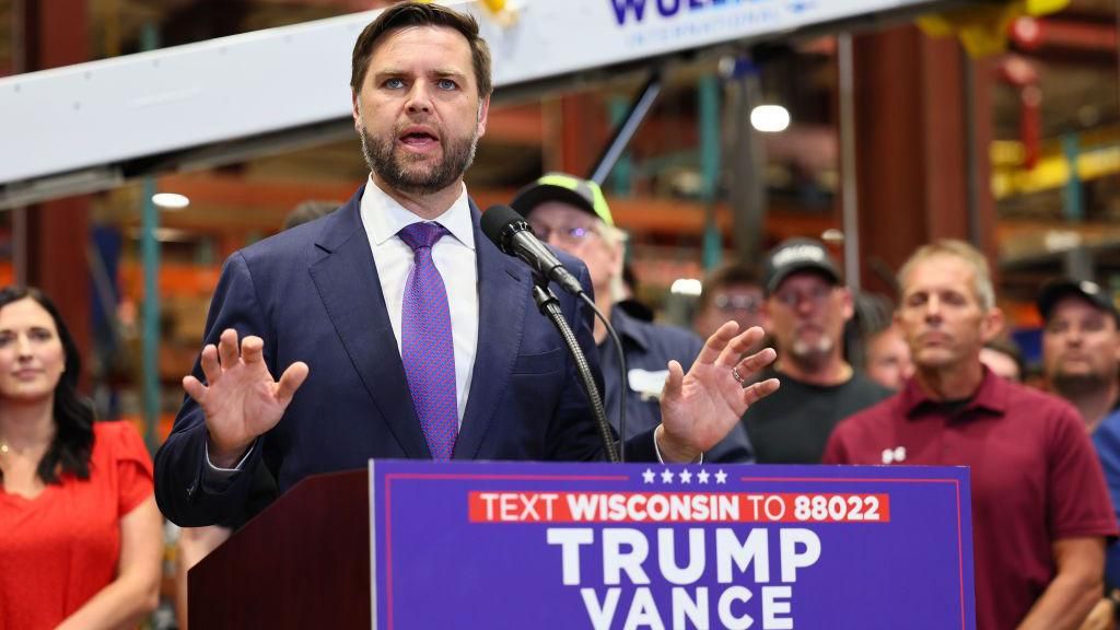 A man with brown hair and a beard, he is wearing a suit with a purple tie standing behind a podium that reads, Text Wisconsin to 88022, TRUMP VANCE