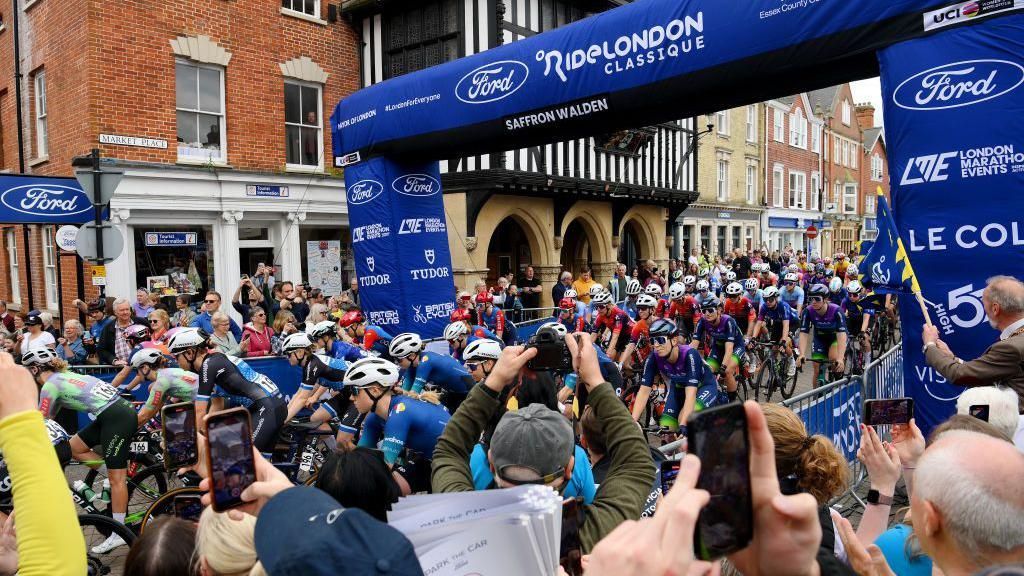 A general view of the peloton prior to the 10th Ford RideLondon Classique 2024 passing under a temporary race gate erected in Market Place, Saffron Walden, with a crowds on either side of the riders behind barriers, and a black and white Tudor-style building in the background