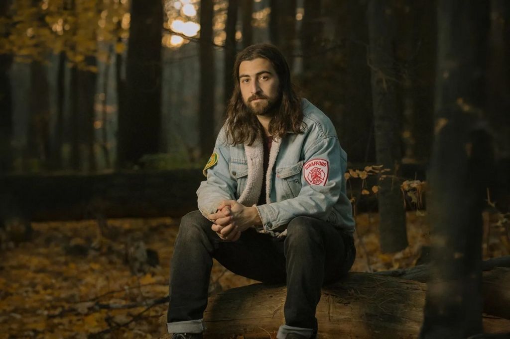 Noah Kahan sits on a fallen tree trunk in a woodland scene. He is wearing a denim jacket and making eye contact with the camera