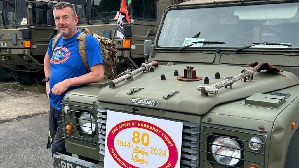 Dave Westall standing beside an army vehicle with a sign for the Spirit of Normandy Trust