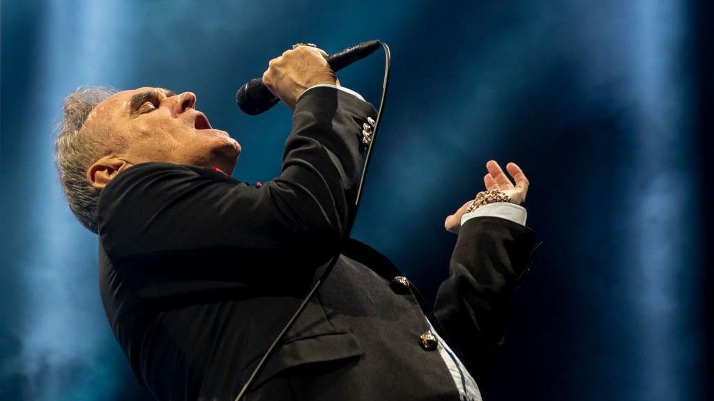 Morrissey bends back as he sings into a microphone, wearing a black suit and tie with a white shirt.