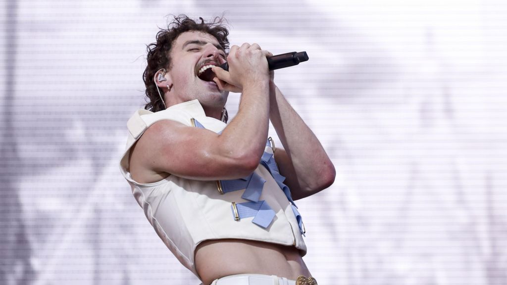 Benson Boone performs on stage. He is holding a microphone in both hands.