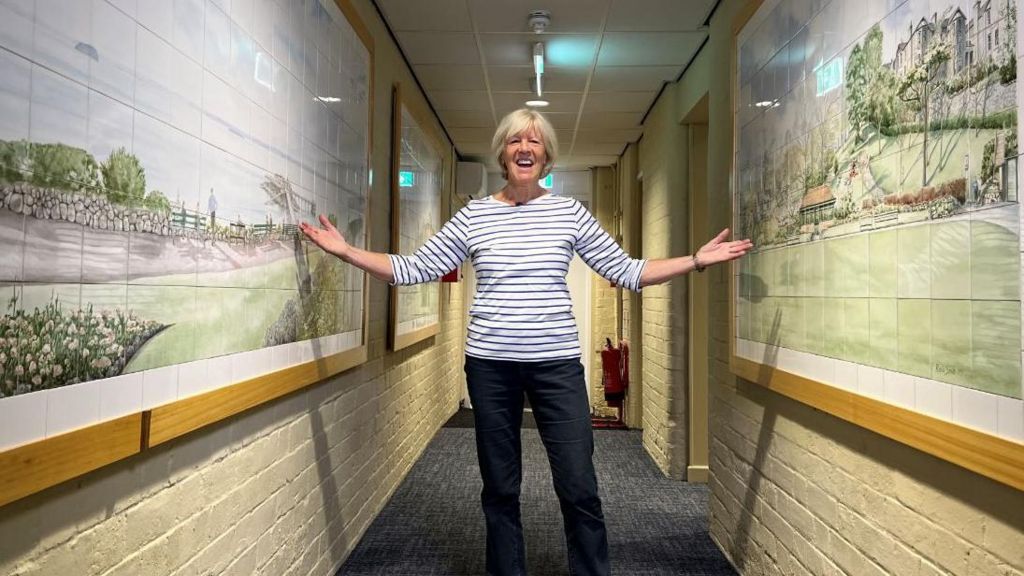 Rosie Smith standing in the corridor with her arms outstretched, gesturing towards her tile artwork displayed on either side of her. She is wearing a black and white striped shirt and black trousers and is smiling. 