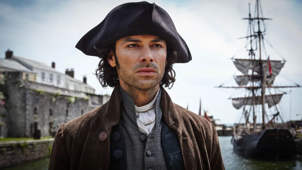 Ross Poldark is looking serious in a harbour which has a large old galleon with its sails furled. There is a stone building to his right. He is wearing a tricorn hat and heavy overcoat over a jacket and a white cravat.