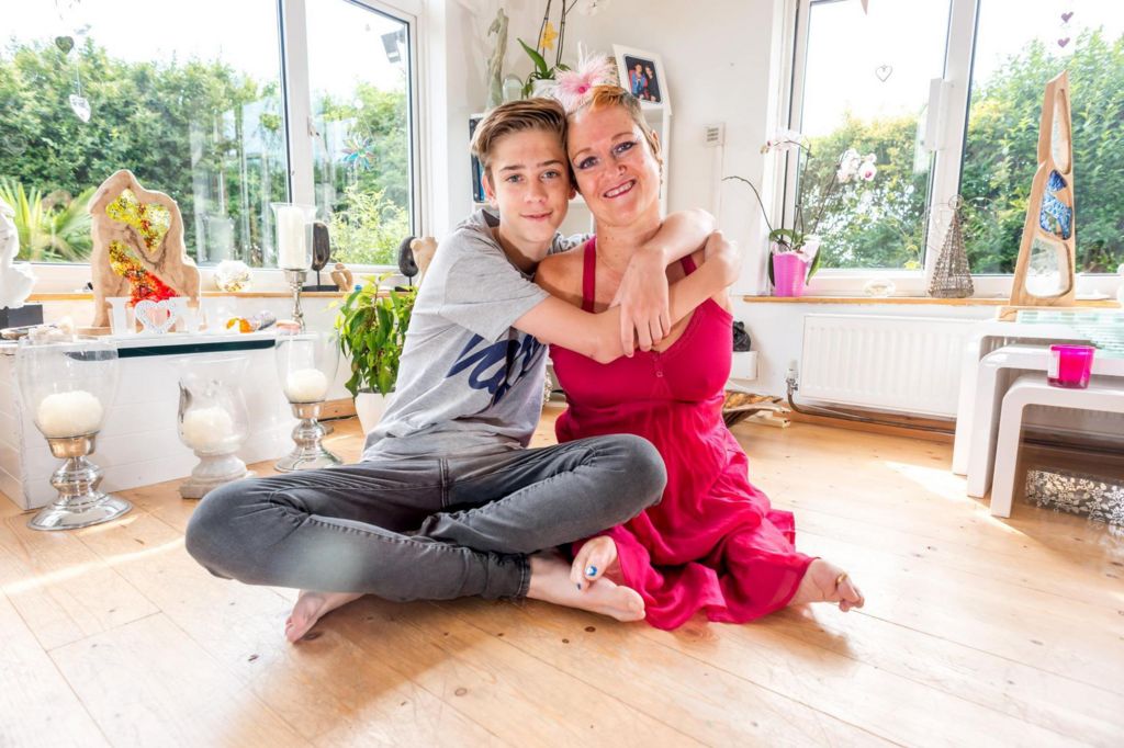 Alison Lapper cuddles her son Parys at their home in 2014. Alison is wearing a pink dress and a streak of her blonde hair is dyed pink. Parys is wearing a grey t-shirt and grey jeans. Artwork can be seen in the room behind them.