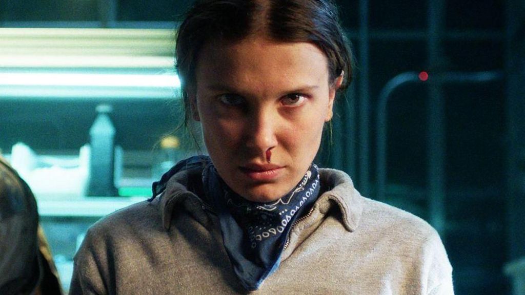 Stranger Things character Eleven, a teenager girl, is stood glaring directly at the camera. She is wearing a grey jumper with a navy, patterned neckerchief visible around her neck. A drip of blood is running from her nose towards her lip. Her head is lowered with her dark hair tied behind her head. Behind her, metallic shelves are visible, lit by neon lights and stacked with what appears to be bottles.