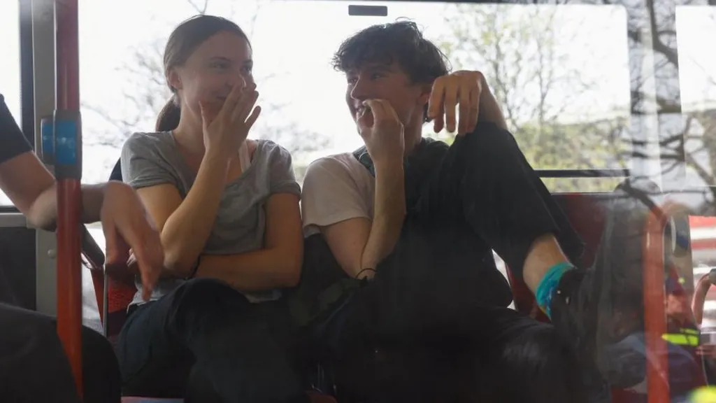 The image shows two young people sitting inside what appears to be a bus or vehicle, both covering their faces with their hands. The person on the left wears a gray t-shirt, while the person on the right wears a light-colored shirt and has curly dark hair. They appear to be seated on red seats, with windows visible in the background showing trees and urban scenery outside.