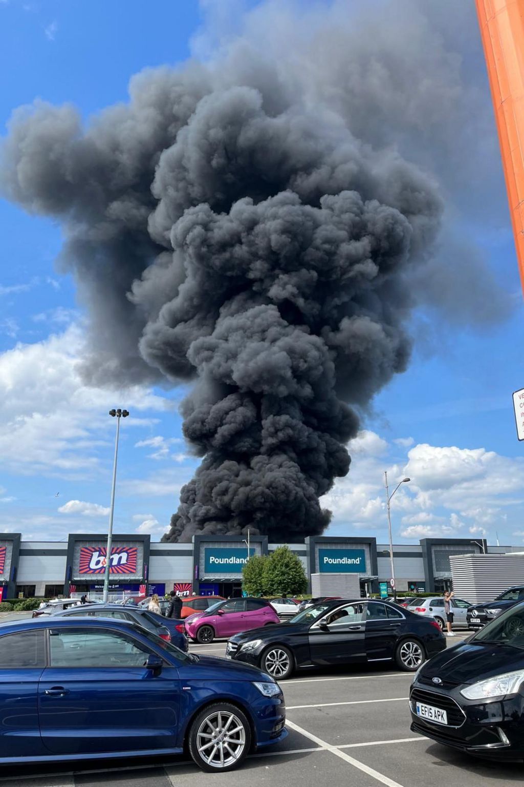 Black smoke plumes rise into a blue sky, above large B&M and Poundland stores, from across a busy car park