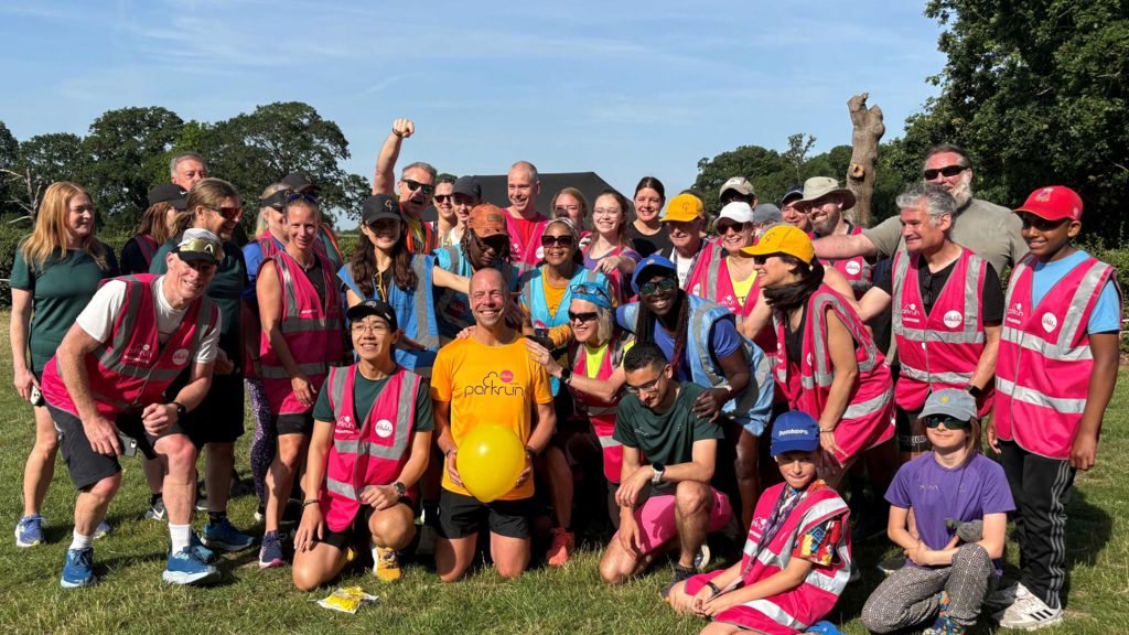 Wood wearing a yellow T-shirt with the word 'parkrun' printed across the front, holding a yellow balloon, surrounded by about two dozen people, most of whom are wearing sun hats, sunglasses and high visibility vests and smiling into the camera.