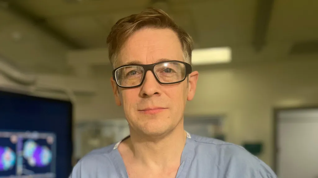 Dr Nick Cromie is wearing light blue scrubs in hospital. He has short light hair and is wearing glasses. 