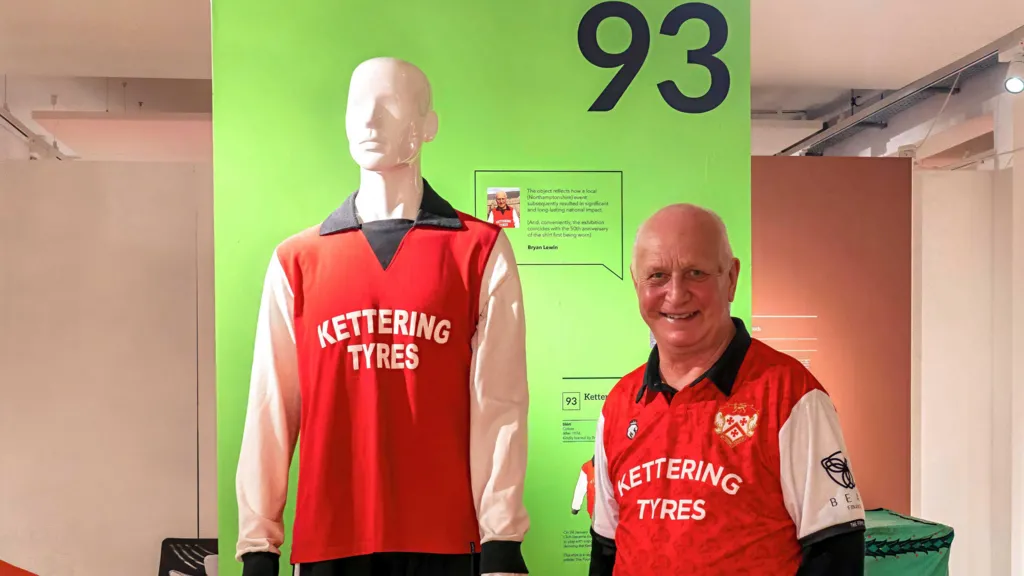 A man at the exhibition standing next to the Kettering Town shirt. The man has short grey hair. 