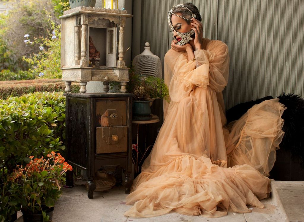 A drag queen in a flowing tuille dress sits next to ruined antique furniture. She has a diamond and pearl encrusted mask around her mouth and draped over her head