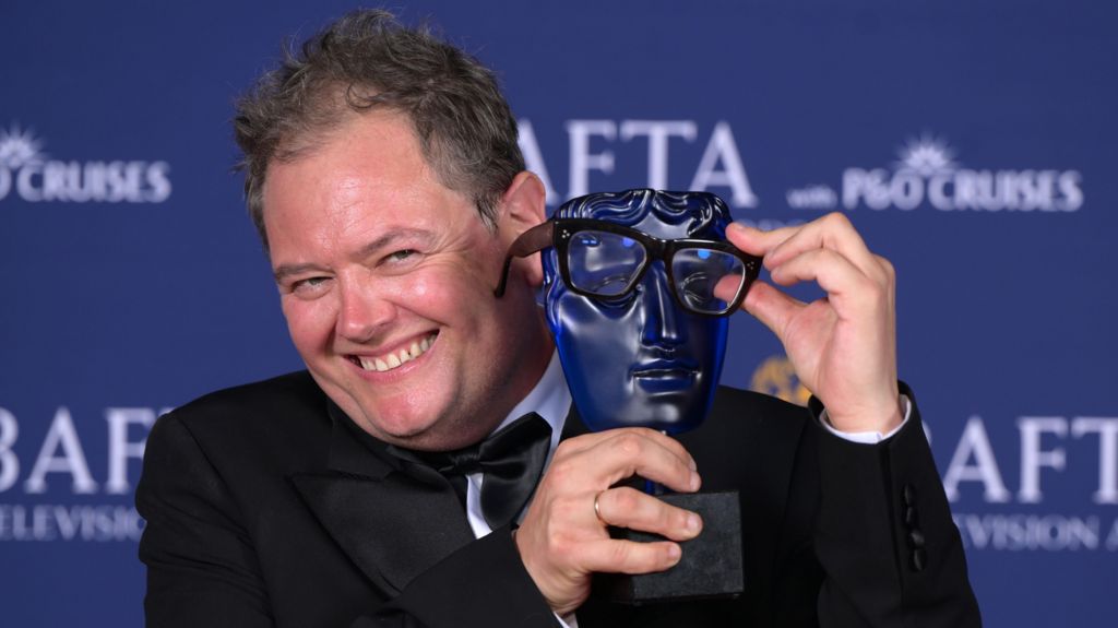 Alan Carr smiling and holding his Bafta face mask trophy, holding his glasses in front of its eyes