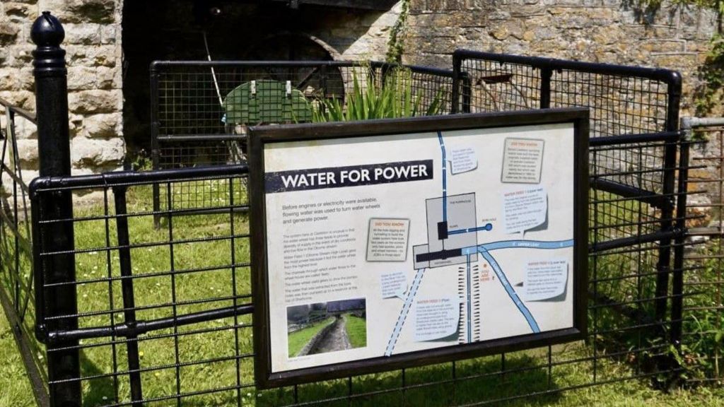 A black wire fence surrounding a water wheel and stone wall. On the fence is an information board with a diagram. The title reads, water for power.