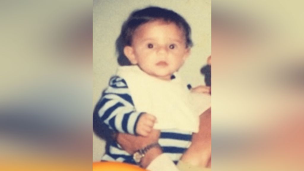 Oliver Archer as a young child. He wears a striped top and a white bib. 