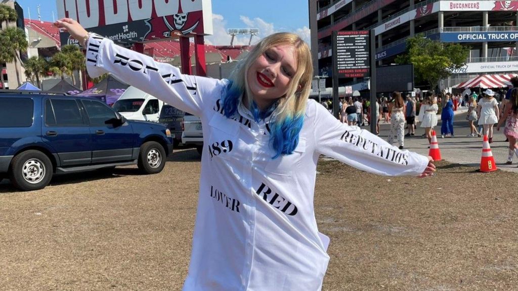 Cali with her arms spread in a homemade white dress with Taylor Swift album names on 