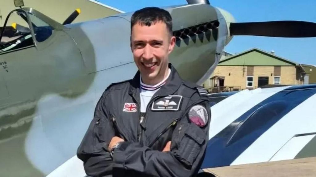 A smiling Sqn Ldr Mark Long, arms folded and wearing a flight suit, standing in front of a Spitfire