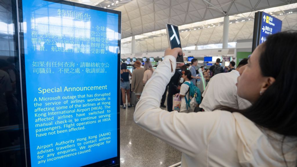 A woman stands in front of a screen which shows a bilingual sign explaining there has been a disruption to airlines worldwide