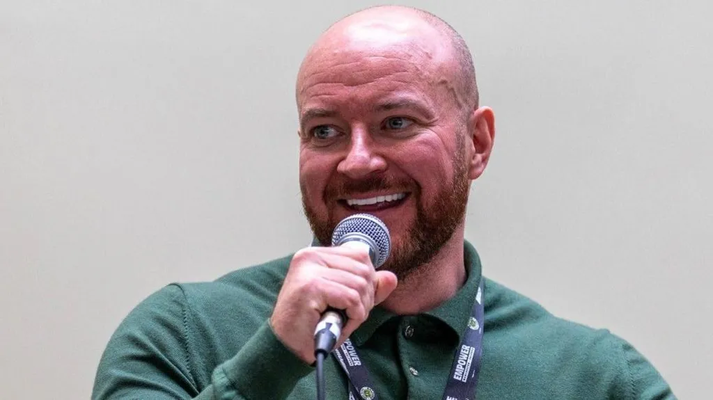A bald man with a dark beard smiles as he clutches a microphone. He is wearing lanyard over a green knit polo. 