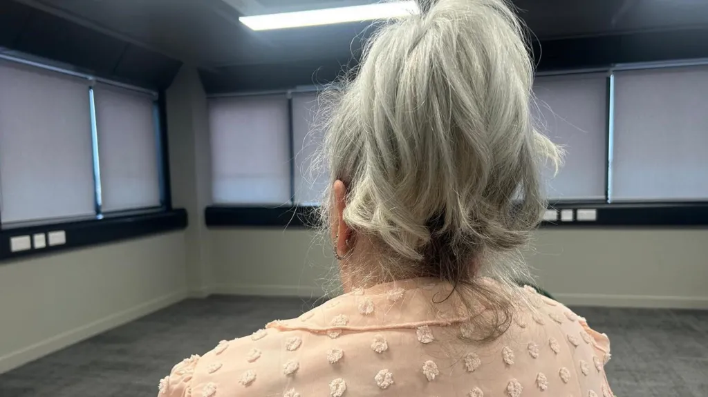 A woman is sitting in a chair with her back to the camera. Her hair is ash blonde and tied up and she's wearing a pink jacket. She's in a bare room with the blinds drawn