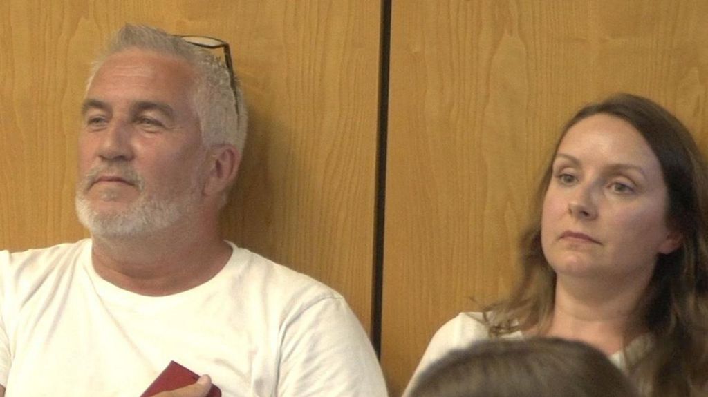 Great British Bake off star Paul Hollywood and his wife Melissa lean against a wood panelled wall in the audience of a council planning meeting. Paul is wearing spectacles on his head.