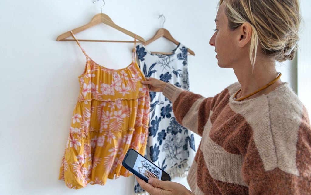 A woman adjusts a dress hanging on a wall in her home, while she holds her phone with the camera app open