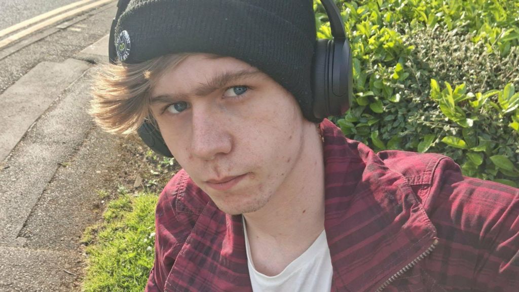 A close-up image of a young man's face. He is frowning, wearing a close-fitting black woollen hat, headphones and a red zip-jacket over a white t-shirt.