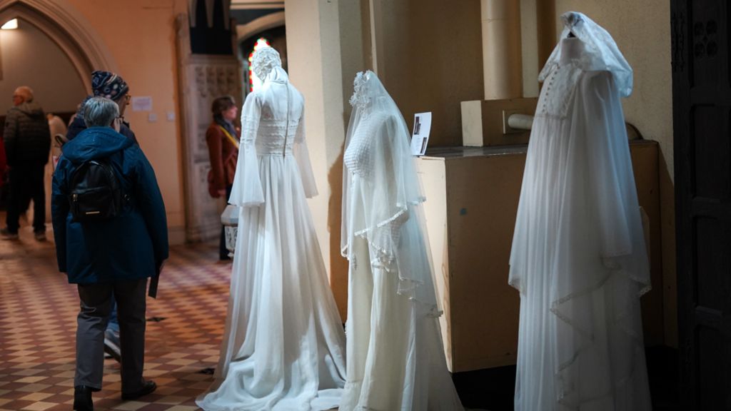Three wedding dresses with two women looking at them and some other dresses in the distance 