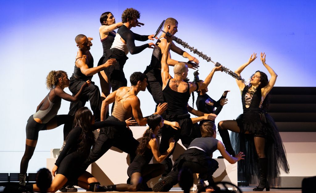 Rosalia on stage posing with her arms in the air, facing 11 dancers who are pointing a long stick or brush at her