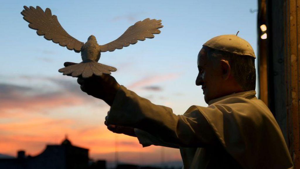 Pope Francis poses with a model of a dove of peace. He holds his arm out and the model sits on his hand.