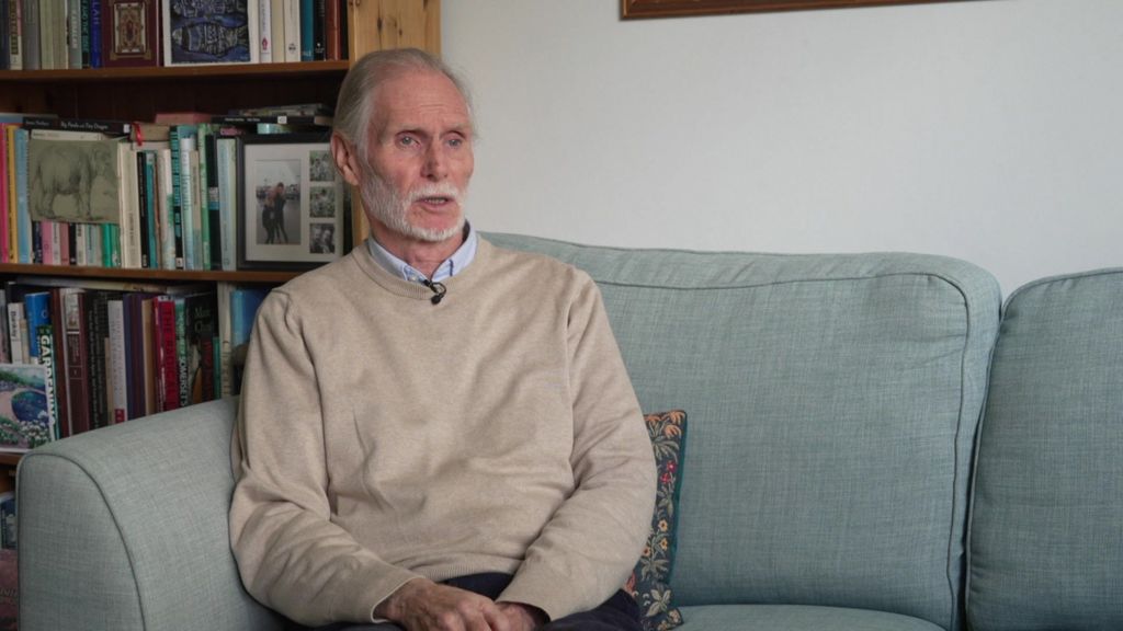 Jim with white hair and a white beard, wearing a beige jumper over a blue collared shirt, sits on a pale green sofa in front of a full bookcase as he speaks during an interview