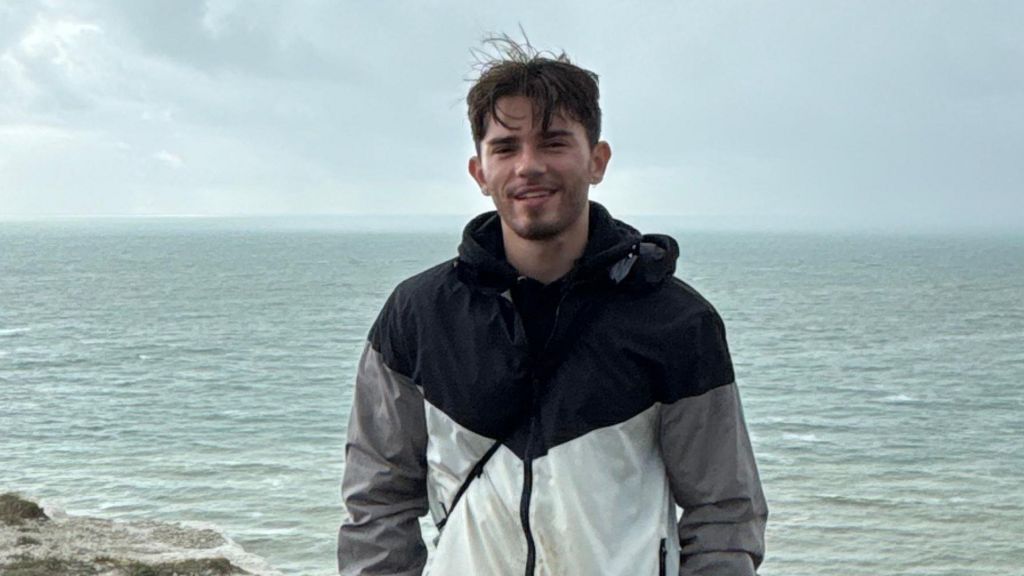 Oliver Archer in a grey, black and white patterned coat, standing in front of the sea. He has dark hair and a beard. 