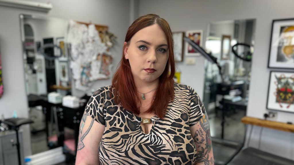 A woman with tattoos on both arms stands in front of tattoo equipment.