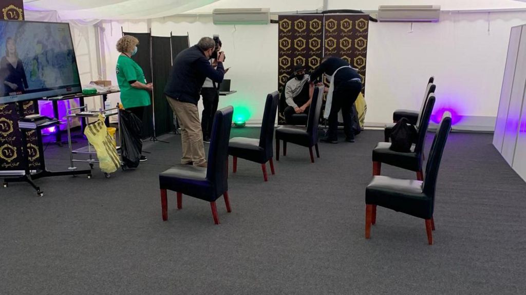 The waiting room for vaccinations with chairs socially distanced and a man receiving his jab in the background