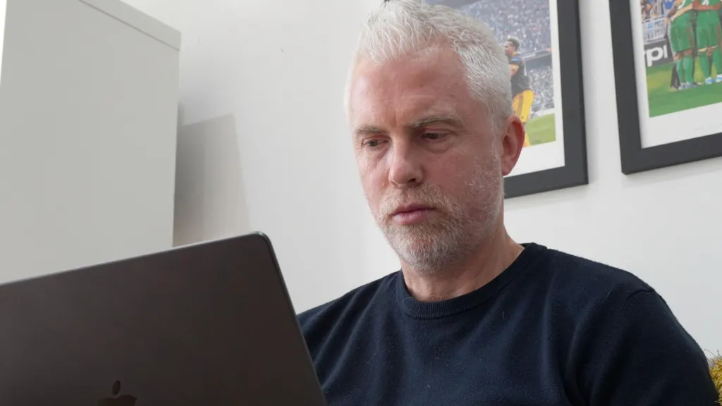 Chris Cobbold who is sitting down and looking at a laptop screen. He has short white hair and a short white beard and wearing a dark blue sweatshirt. 