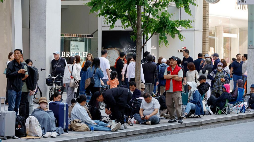 Scores of people sit and stand as they wait outside a store with various watch firms branded on a store window. There is tree inthe middle of them.