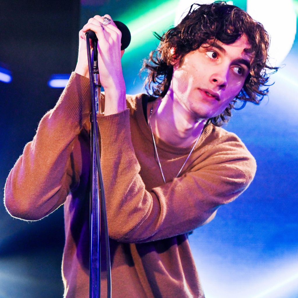 Sombr wearing a brown jumper holding a microphone stand with both hands and looking off camera
