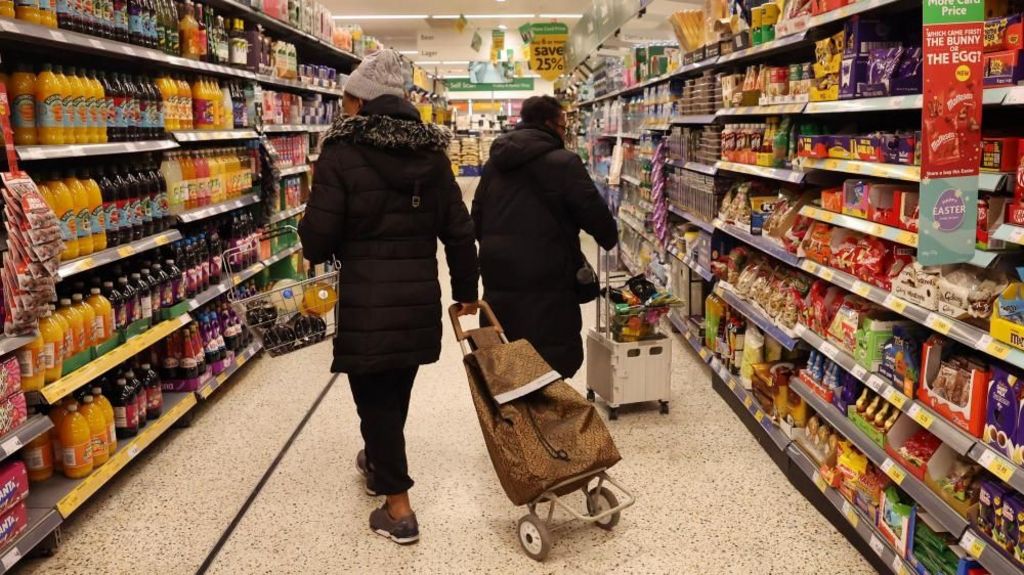 Customers holding a basket and wheeling a trolley bag shop walk down a supermarket aisle.